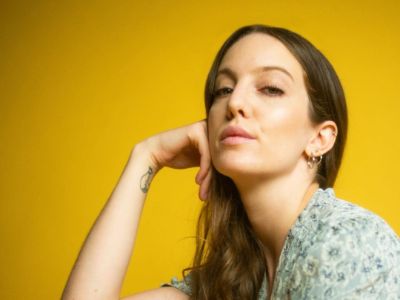 Eleanor Lambert is posing by putting her right hand on her right cheek in front of a yellow background.